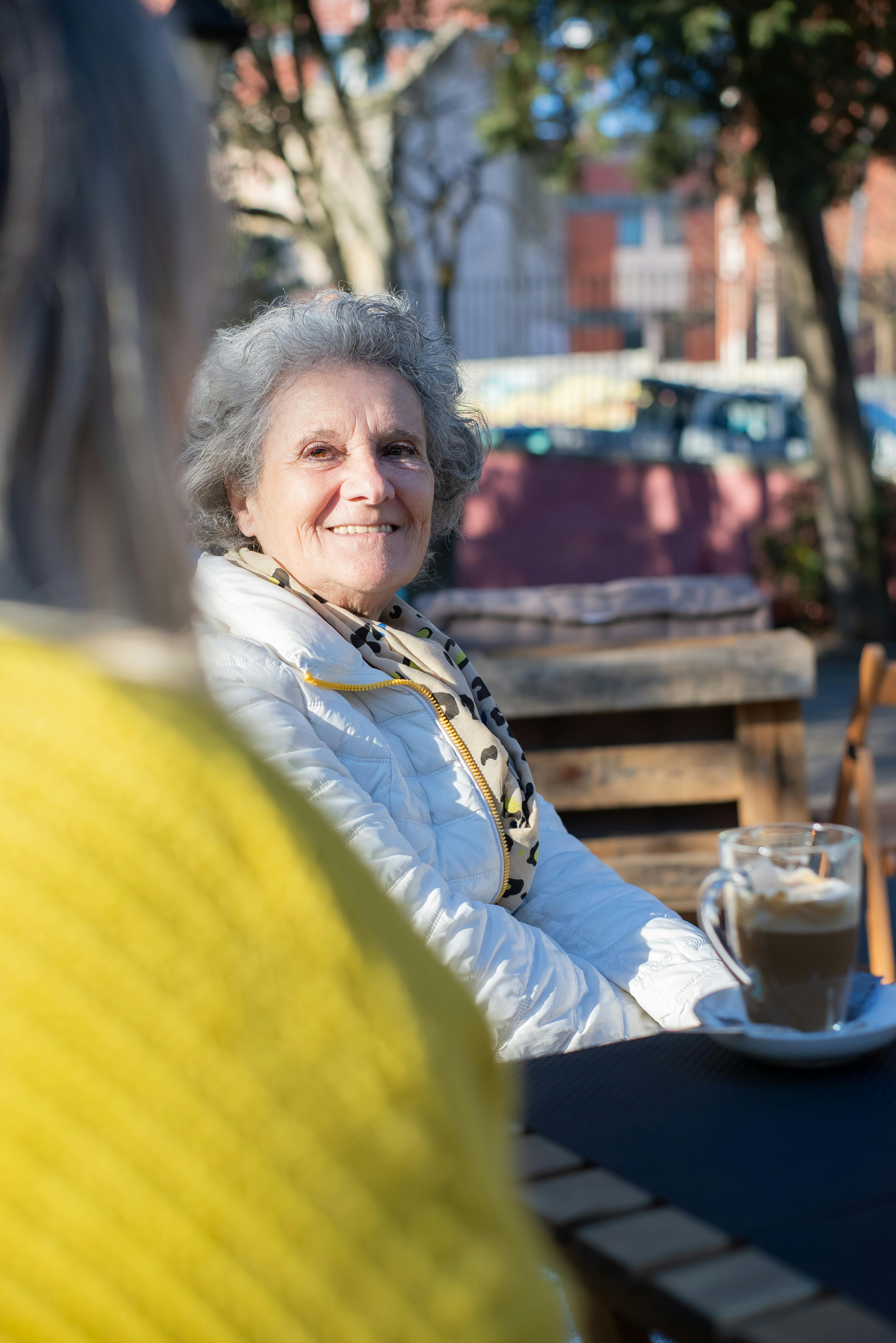 An older woman smiling while having coffee outdoors with a caregiver